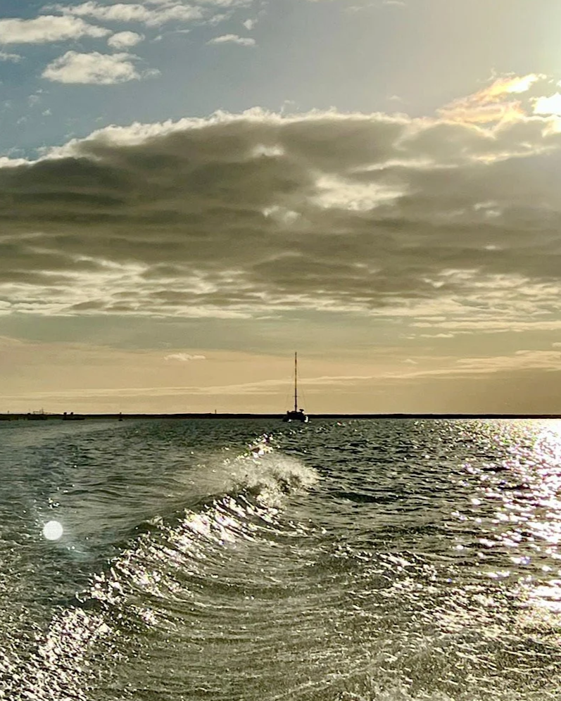 The wake of a boat crossing the Ria Formosa near Ilha da Culatra, a short ferry from Casa Fuzetta, with a distant sailboat on the horizon under a low afternoon sun.