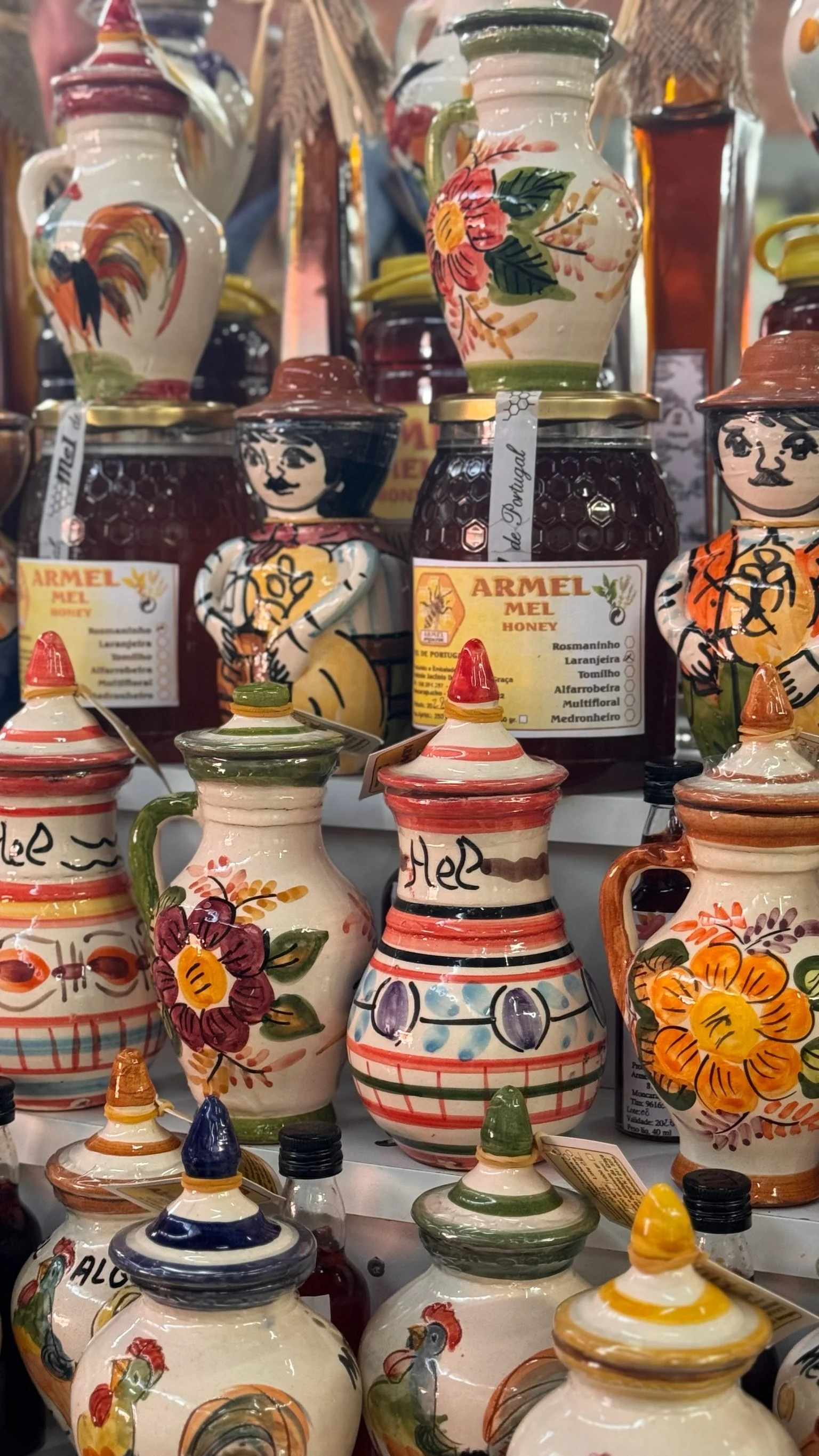A market stall in Olhão close to Casa Fuzetta stacked with hand-painted Portuguese ceramic honey jugs in floral and folk-art designs, alongside jars of Armel honey.