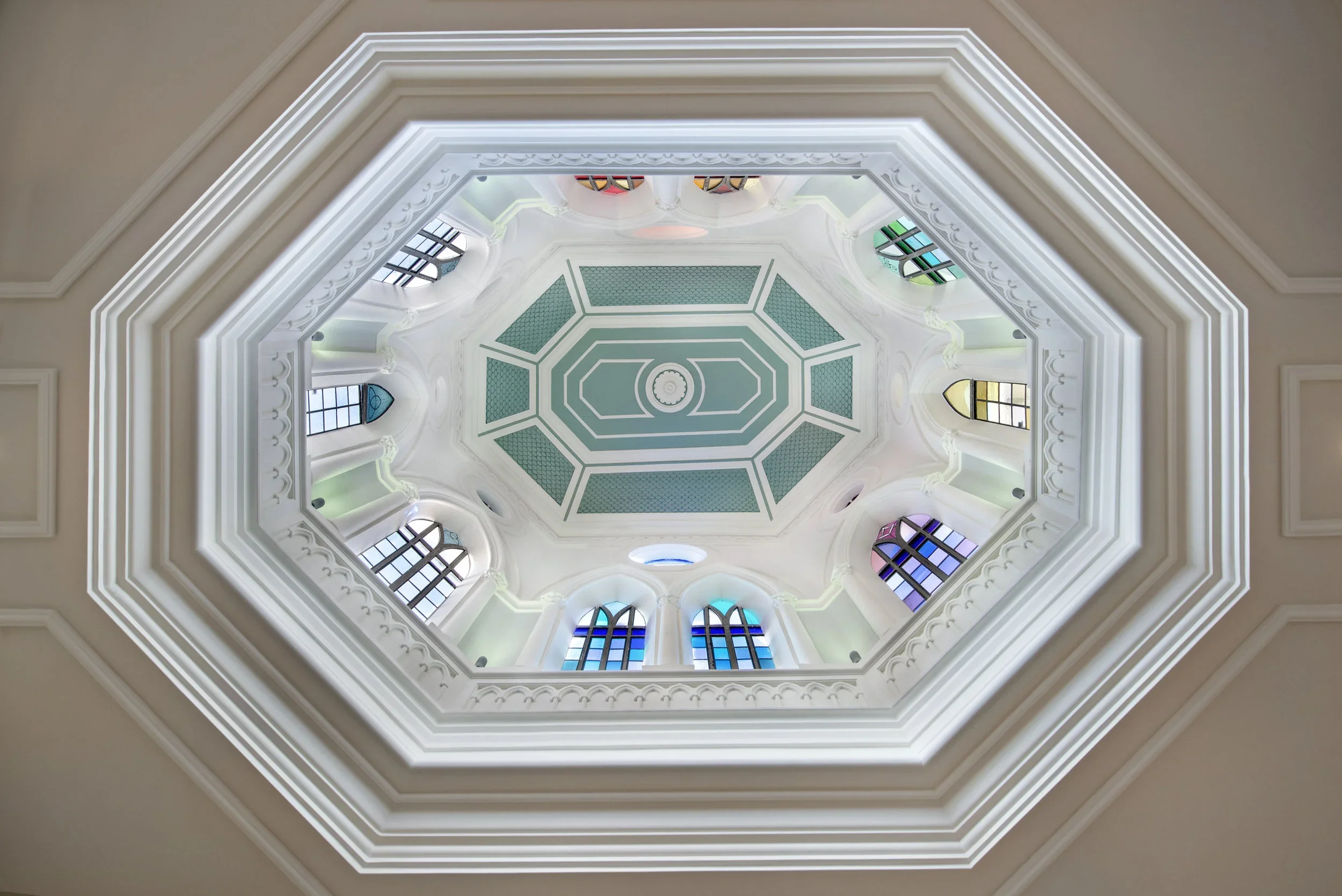 The domed ceiling of the meditation room at Casa Fuzetta — an octagonal cupola with ornate white plasterwork, a pale green coffered centre and arched stained-glass windows in jewel tones.