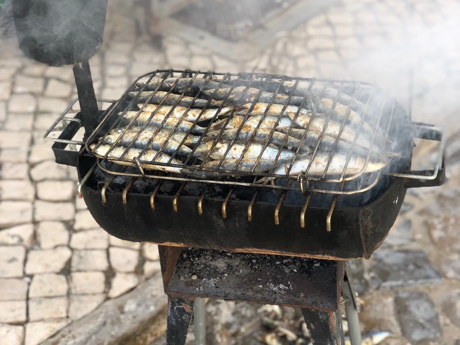 Fresh sardines grilling over hot coals on a street-side barbecue on the hand-cut calçada cobblestones of Olhão, home to Casa Fuzetta, with smoke rising in the sunlight.