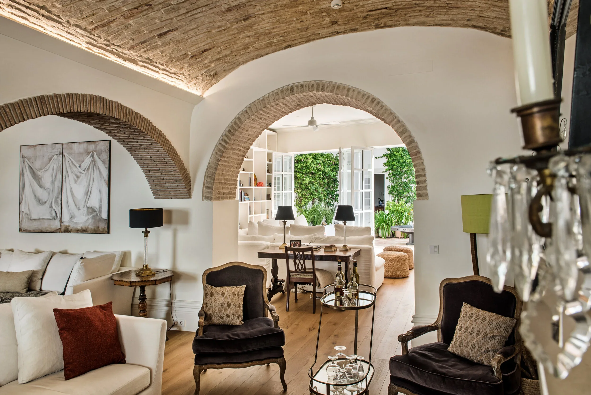 The living room and library at Casa Fuzetta; white linen sofas and velvet armchairs beneath an exposed brick vaulted ceiling and arches, opening through to bookshelves and a green courtyard.