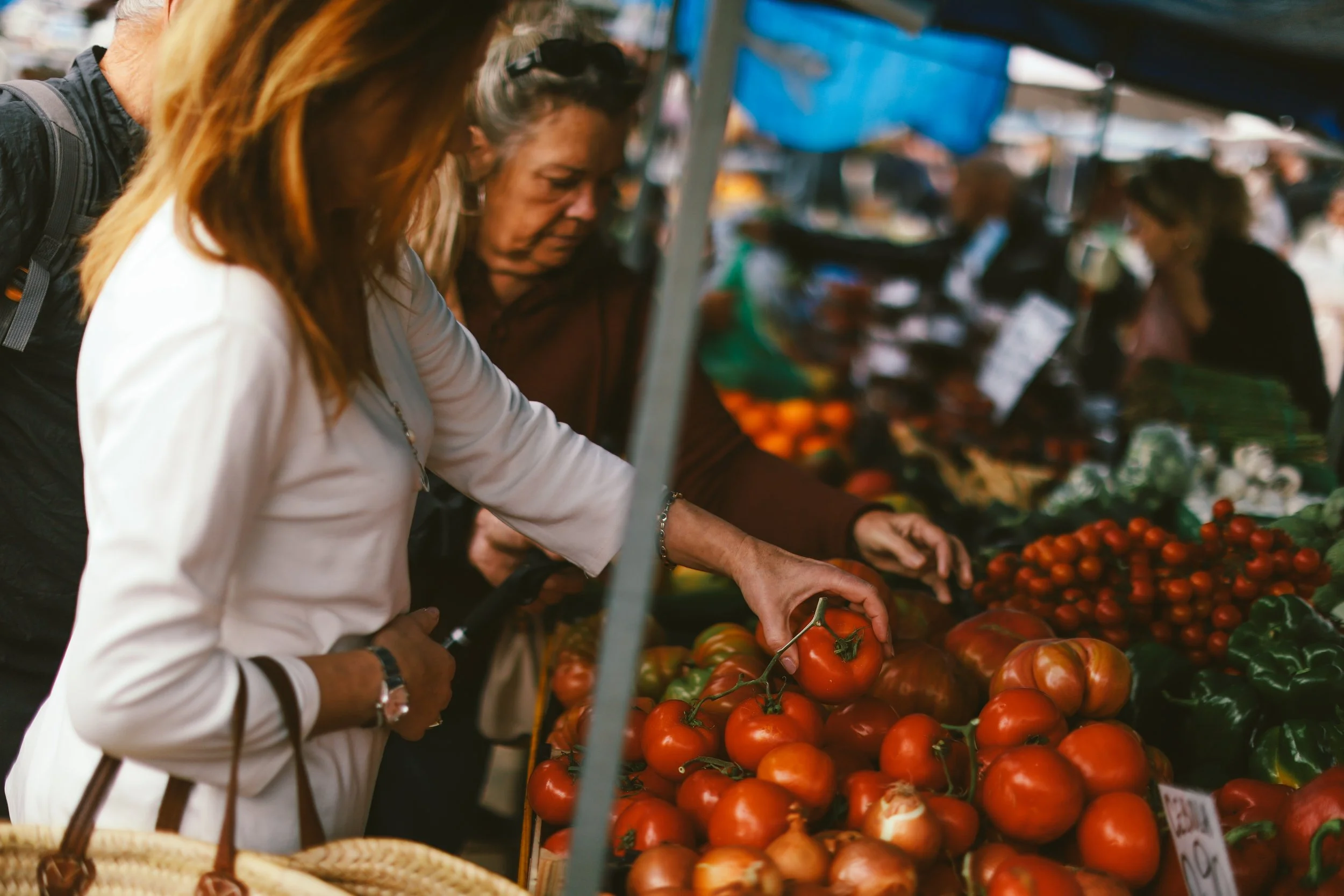 A shopper selects vine tomatoes at a produce stall in Olhão's market, a short walk from Casa Fuzetta, with crates of oranges, chard and cherry tomatoes under a blue awning.