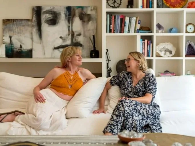 Two Cheeky Yoga Club retreat guests in conversation on a white sofa in a Casa Fuzetta sitting room in Olhão, with a bookshelf holding crystals and abstract portraits on the wall behind.