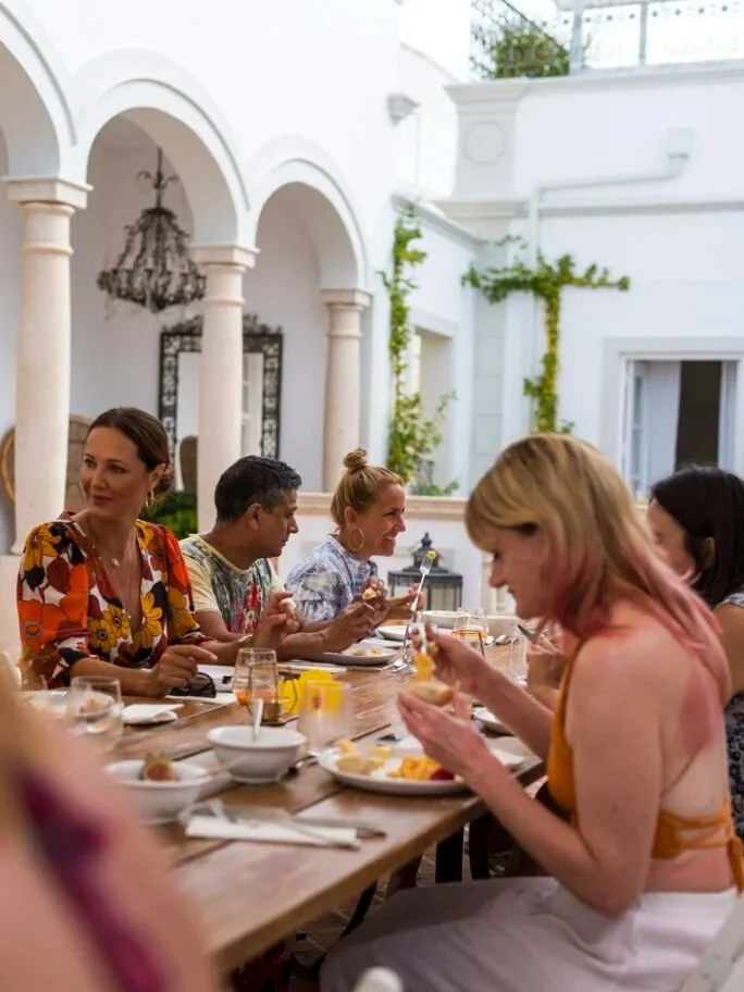 Cheeky Yoga Club retreat guests at a long-table lunch in Casa Fuzetta's colonnaded courtyard in Olhão, under the arched arcade with a wrought-iron chandelier and climbing greenery.