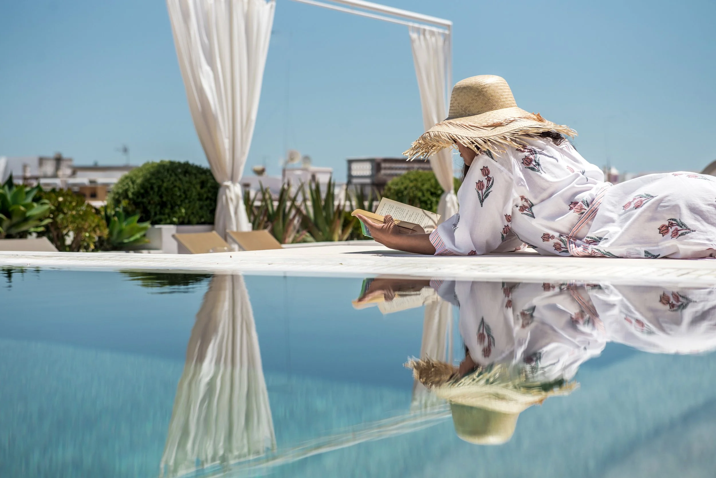 image of a guest wearing a Casa Fuzetta pink tulip robe lying by the pool on the rooftop of Casa Fuzetta reading a book and wearing a straw hat with her reflexion mirroring in the surface of the swimming pool
