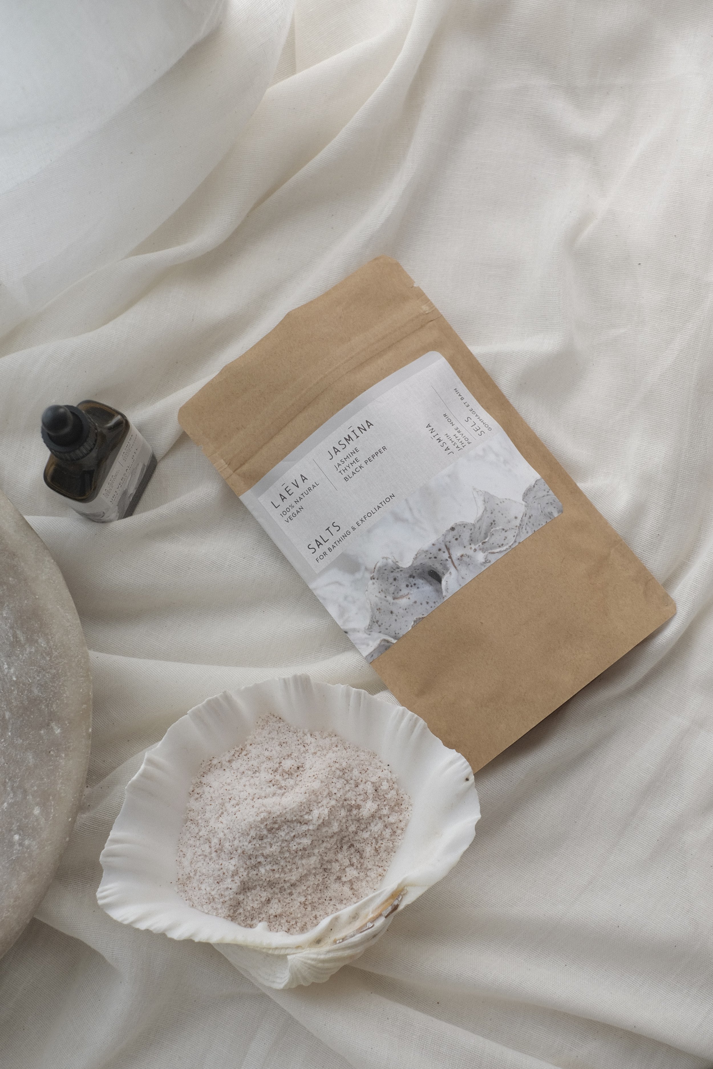 La-Eva Jasmina bath salts in a shell dish beside the matching Jasmina oil, styled on soft white linen