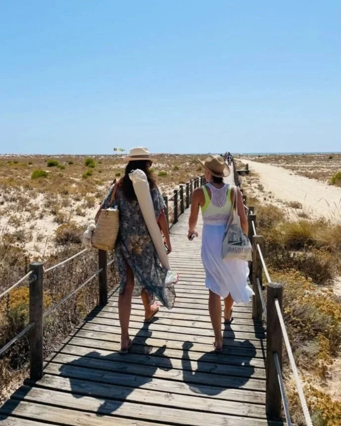 Two Cheeky Yoga Club retreat guests walk the wooden boardwalk to the beach on Ilha da Armona, a short ferry from Casa Fuzetta in Olhão, with beach mats and sun hats under a bright blue sky.