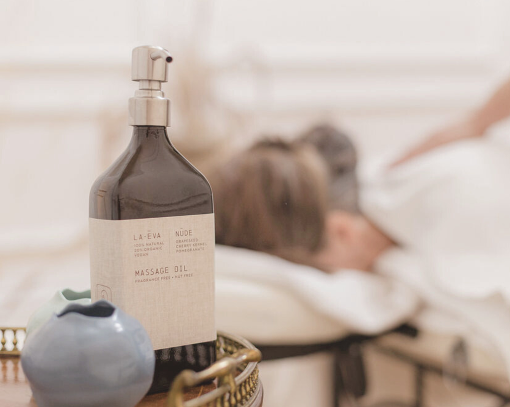A La-Eva Nude Massage Oil bottle — a fragrance-free, nut-free grapeseed, cherry kernel and pomegranate oil — on a brass tray beside a ceramic pot, with a guest on the massage table behind at Casa Fuzetta.