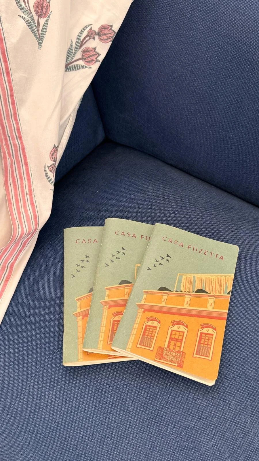 Three exclusive Casa Fuzetta notebooks — sage-green covers illustrated with the house's Olhão façade — fanned on a navy linen chair beside Casa Fuzetta's floral silk robe with its pink-striped border.