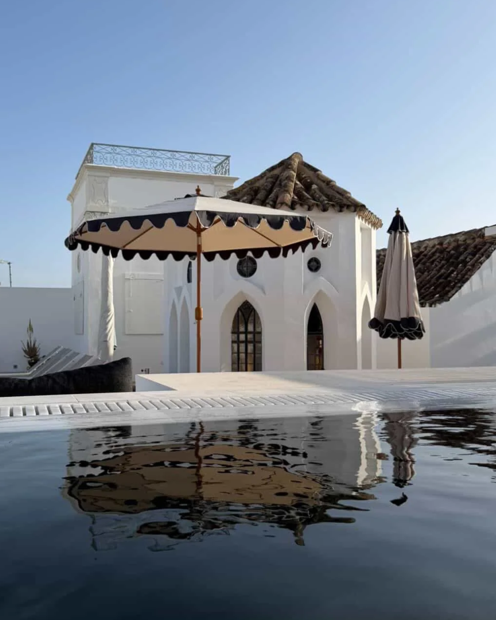 The rooftop pool at Casa Fuzetta in Olhão — dark water reflecting the white Moorish-arched pavilion under a cream-and-navy parasol, with terracotta roofs and clear blue sky beyond.