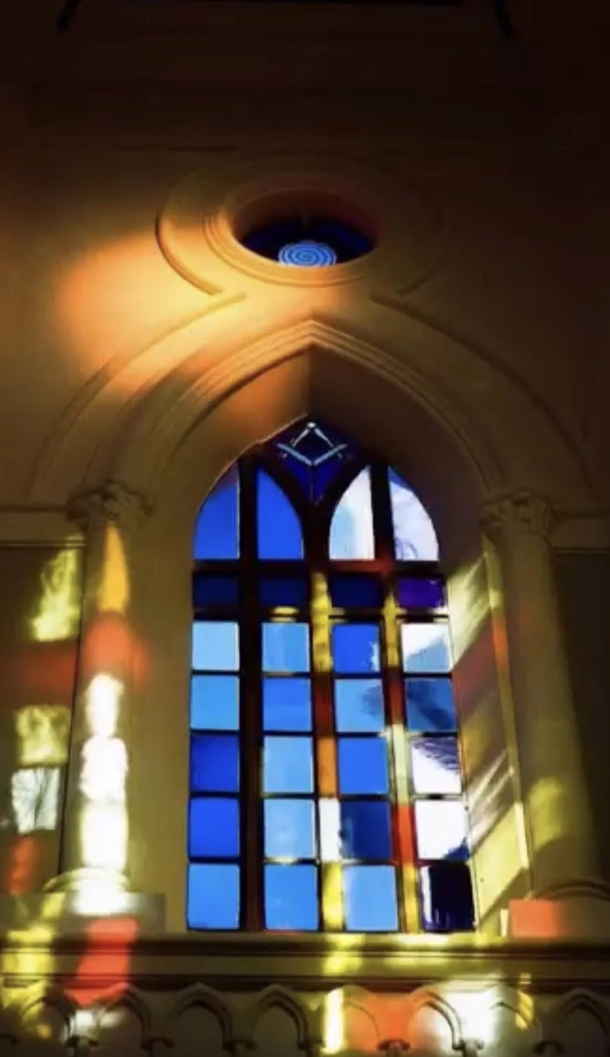 Sunlight streams through a Gothic-arched window of blue stained glass in the meditation room at Casa Fuzetta in Olhao, below a round spiral window, scattering gold and red light on the walls.