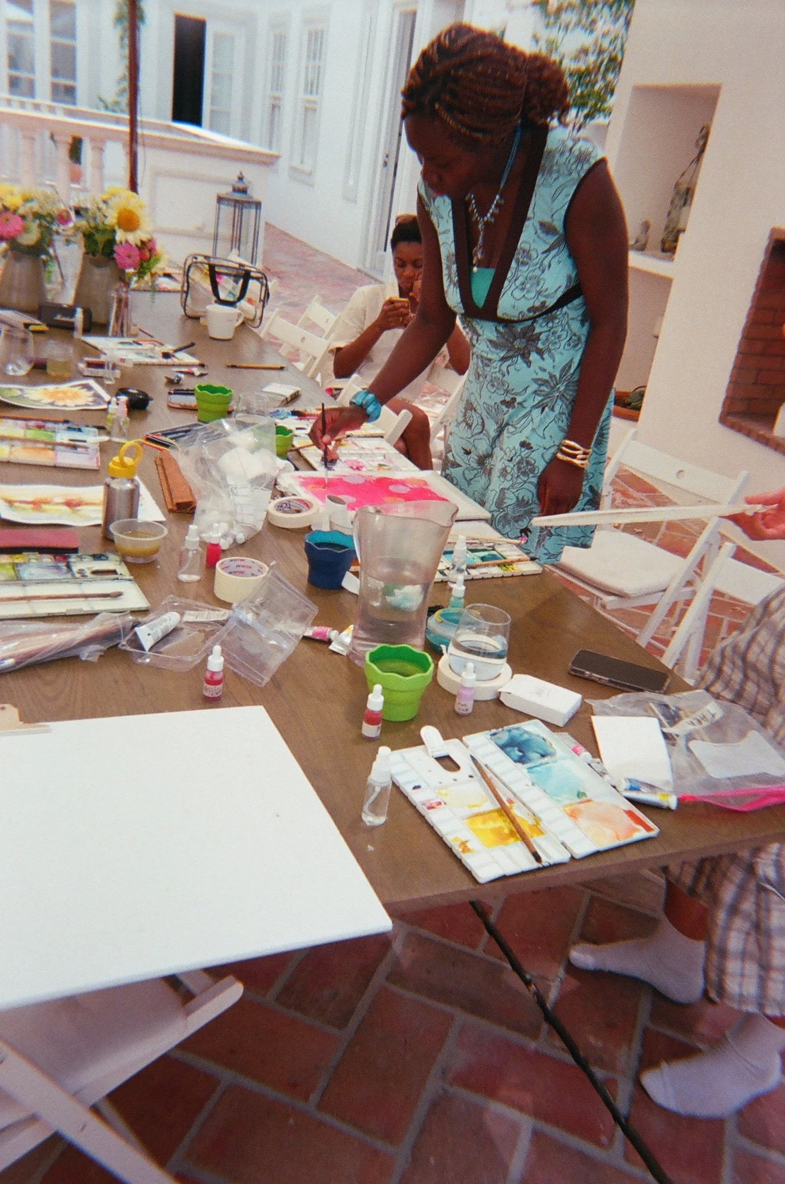 A watercolour class on the kitchen terrace at Casa Fuzetta in Olhao; a painter in a turquoise floral dress lays a bright pink wash at a long table of palettes, brushes and summer flowers.