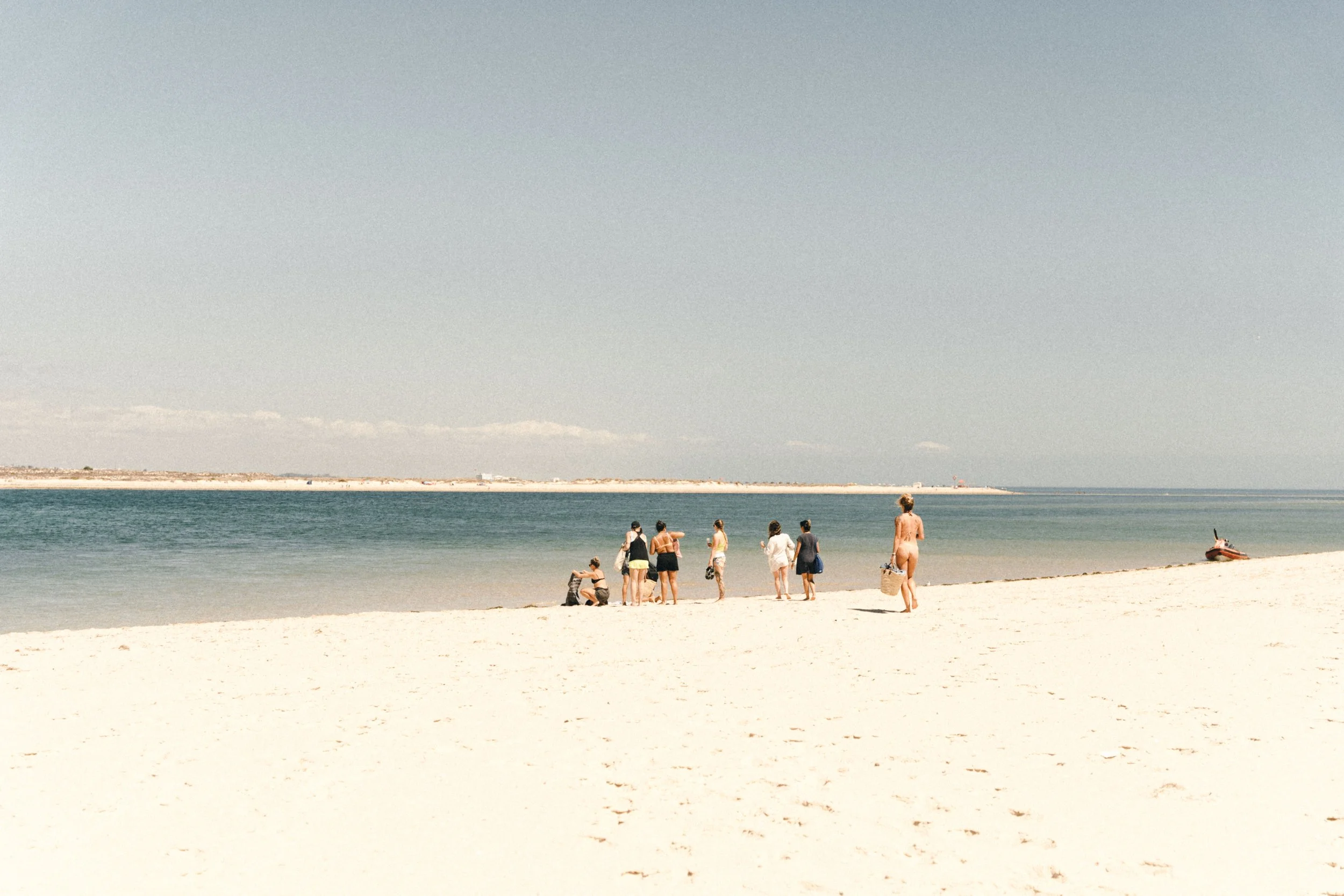 Retreat guests on a white-sand beach on one of the Ria Formosa islands, a short boat ride from Casa Fuzetta in Olhao, gathered at the water’s edge with beach bags and a straw basket.