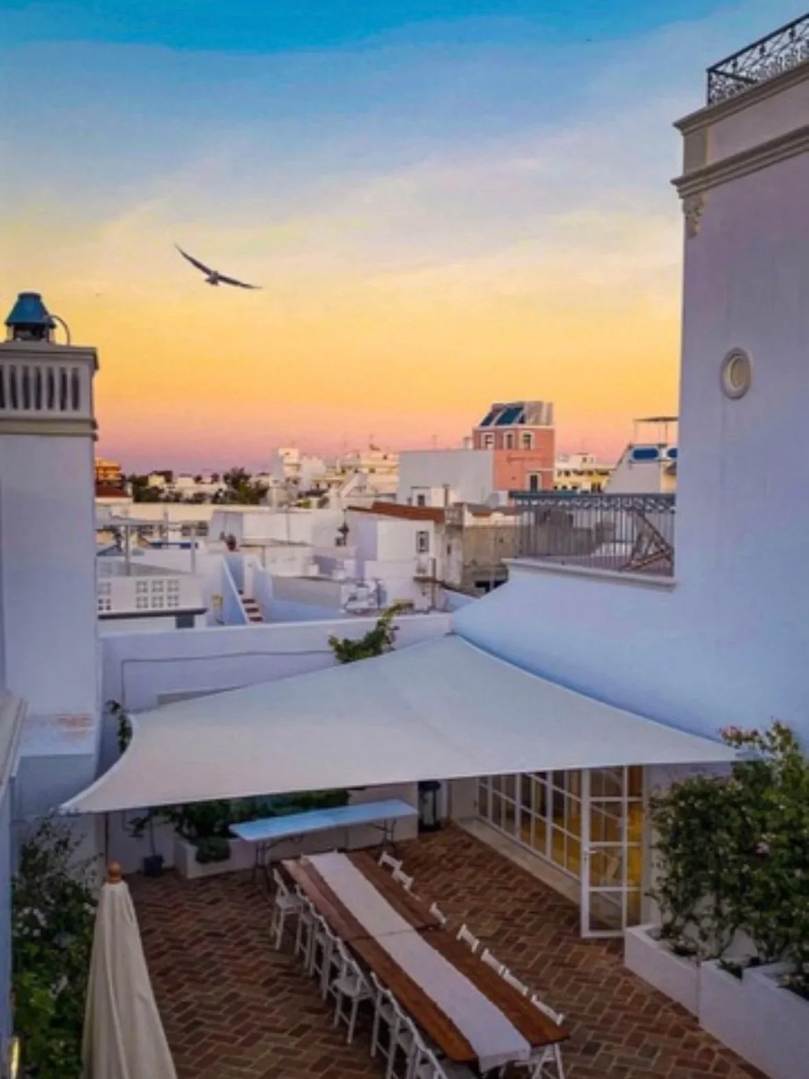 The kitchen terrace at Casa Fuzetta at sunset; a long wooden dining table beneath a white sail canopy on herringbone brick, a gull passing above Olhao's whitewashed rooftops to the horizon.