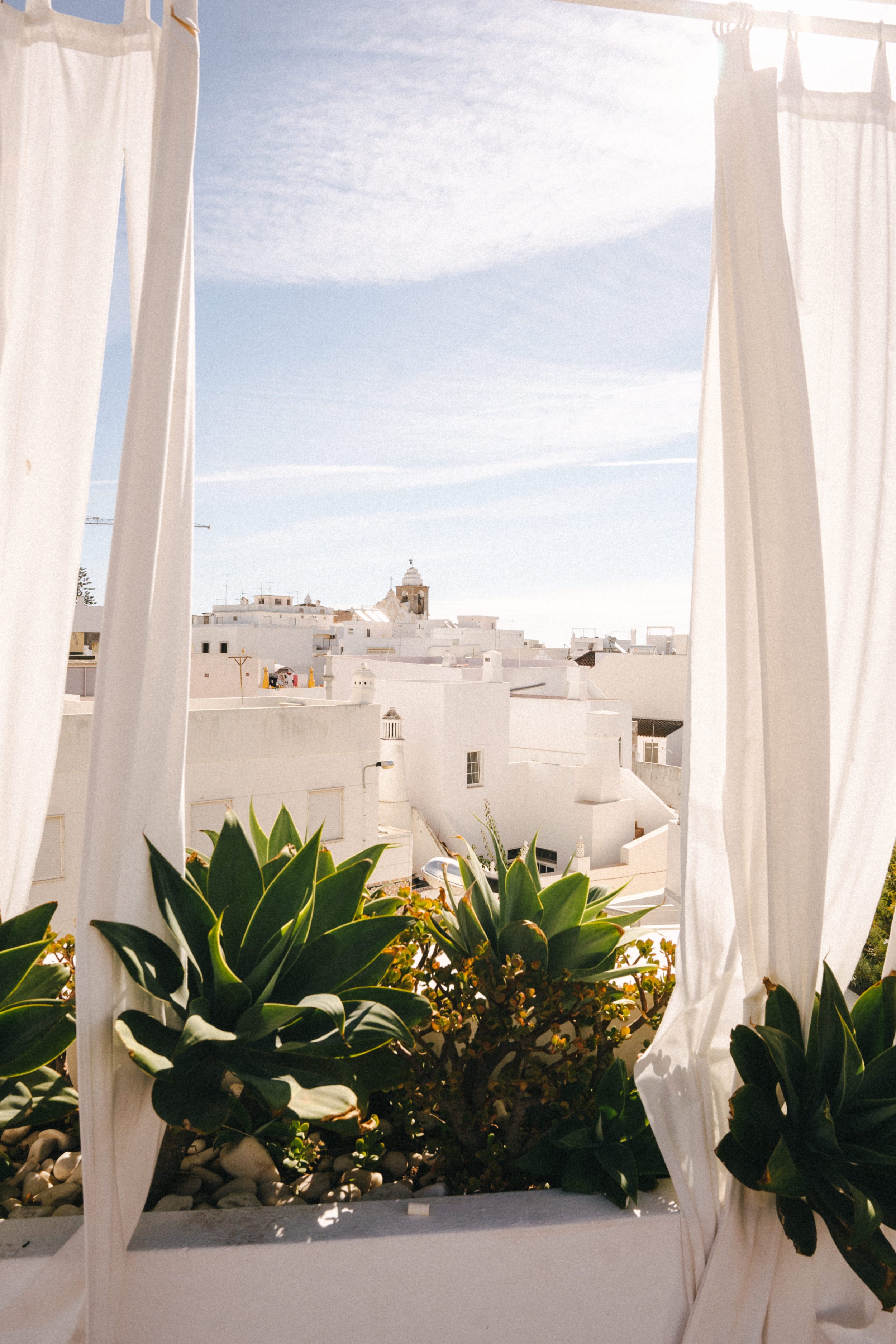 White linen curtains frame a view of Olhão's whitewashed rooftops and church dome from a rooftop terrace at Casa Fuzetta, with succulents in the foreground under a bright sky.