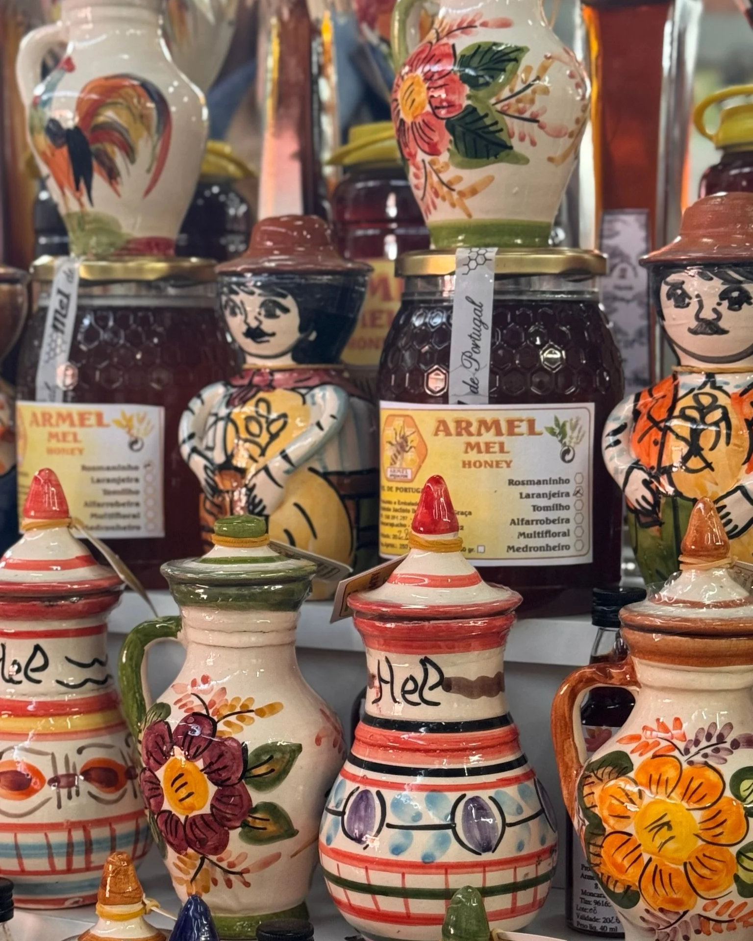 A display of hand-painted Portuguese ceramic honey pots at a market stall in Olhão, close to Casa Fuzetta, with folk-art figures and jars of Armel honey in floral and rooster motifs.