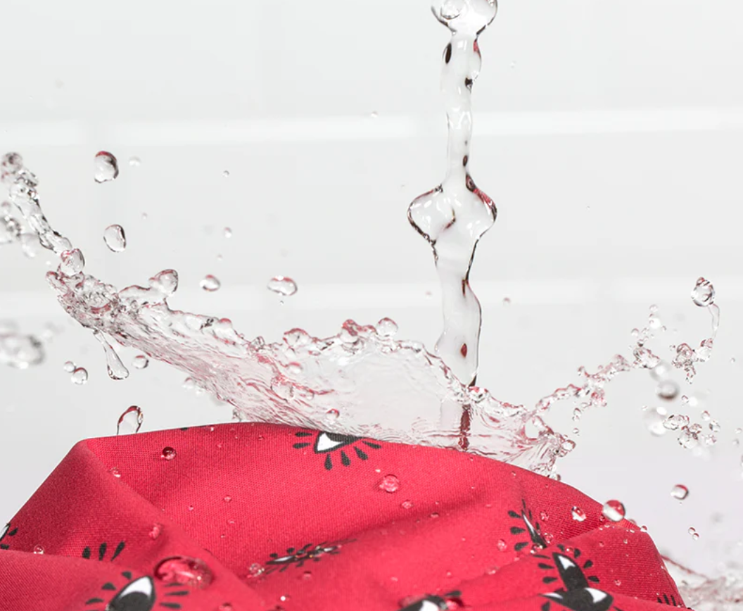 Close-up of a red Shhhowercap turban shower cap printed with small black-and-white eyes, with water splashing off its 100% waterproof surface.