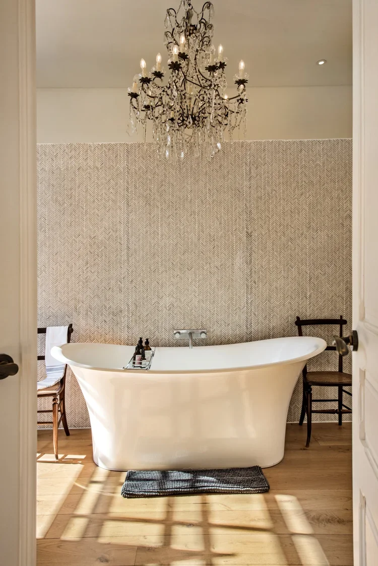 The ensuite bathroom of the Crystal Suite at Casa Fuzetta in Olhao, with a freestanding roll-top bath against a herringbone tiled wall, a beaded crystal chandelier and sunlit oak floors.