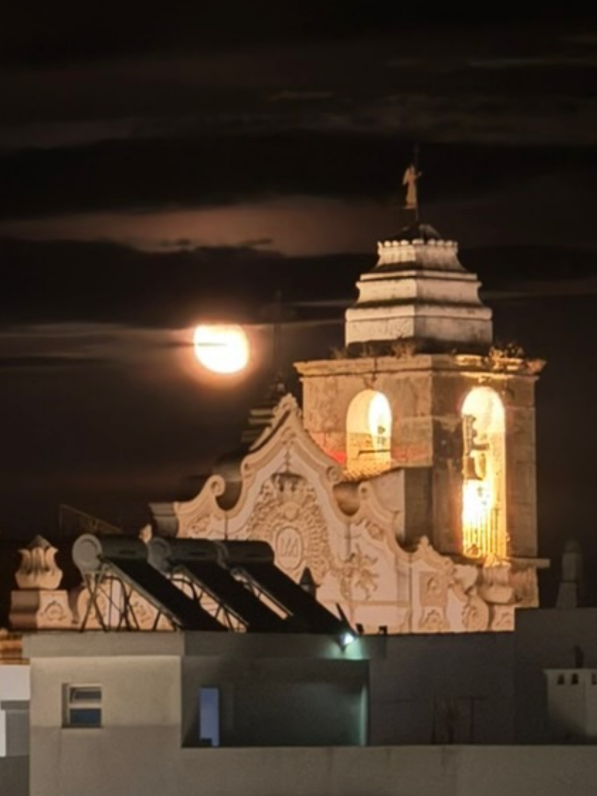 A full moon beside the floodlit bell tower of the Igreja Matriz in Olhao, close to Casa Fuzetta, its scrolled baroque gable glowing white against a dark, cloud-streaked night sky.