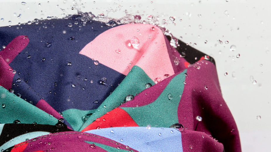 Close-up of a Shhhowercap turban shower cap in a multicoloured abstract print — navy, pink, purple, teal and red — with water beading on its 100% waterproof surface, featured in Elle (US)