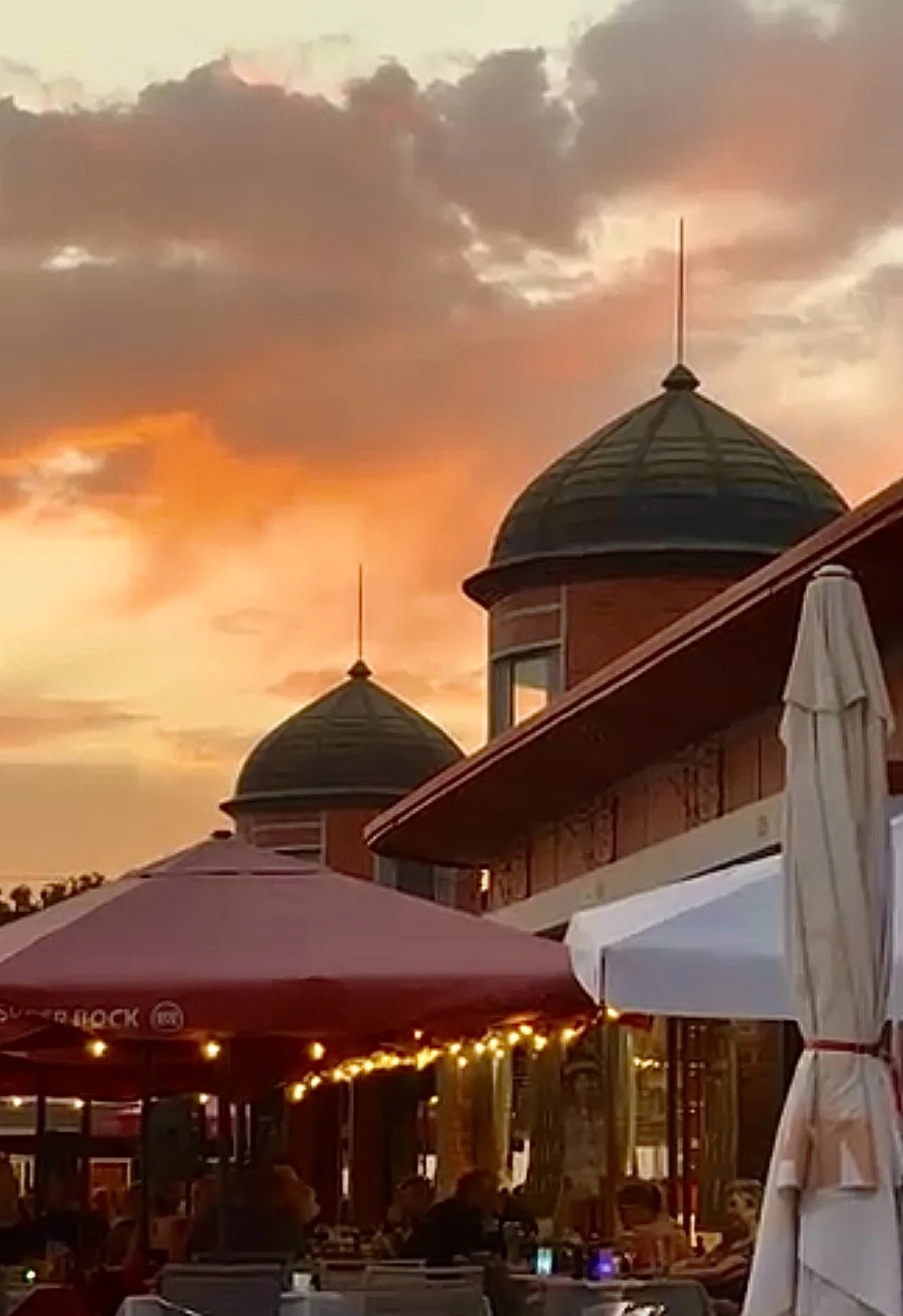 The green domes and red-brick towers of the Olhão market halls against a fiery sunset sky, a short walk from Casa Fuzetta, with café parasols and festoon lights on the terrace below.