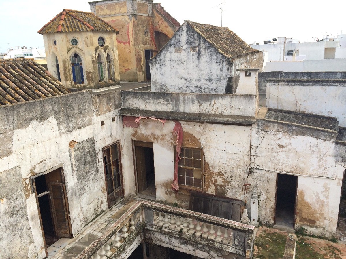 Casa Fuzetta before its restoration — derelict, weather-stained buildings with cracked plaster, broken windows and a crumbling balustrade, a turret of pointed stained-glass windows beyond, in Olhão.