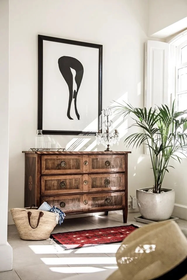 A sunlit corner at Casa Fuzetta — an antique wooden chest of drawers below a framed black-and-white artwork of a woman's legs, with a crystal candelabra, a potted palm and a woven basket.