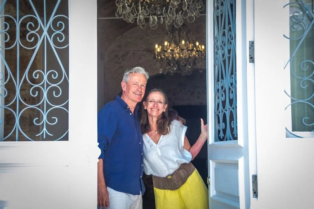 Casa Fuzetta founders Tara Donovan and Jonathan Tod, smiling arm in arm in the grey and blue wrought-iron doorway of the house in Olhão, with crystal chandeliers and a vaulted stone interior behind them.