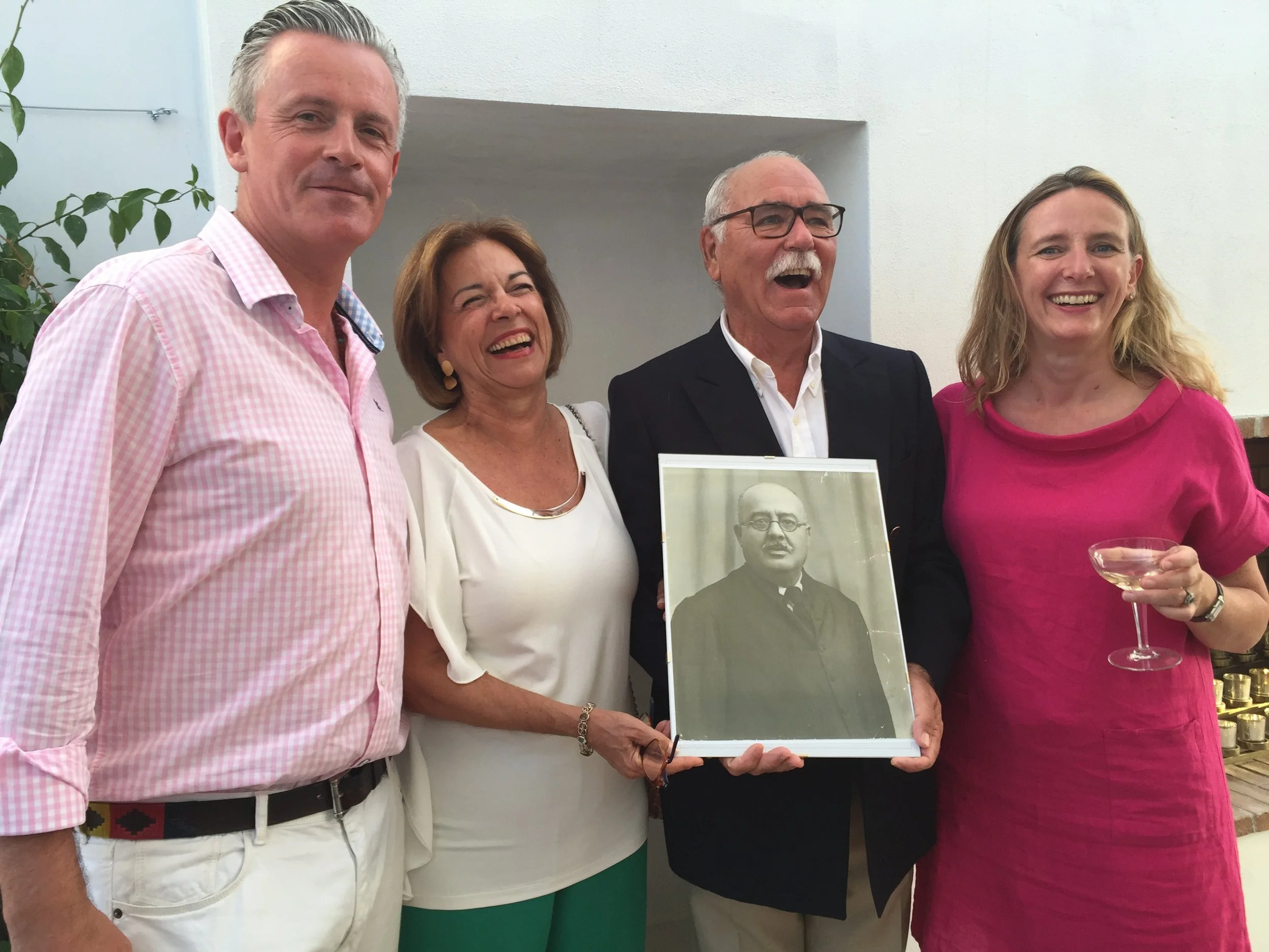 The owners of Casa Fuzetta, Jonathan and Tara, with the grandson of Dr. Carlos Fuzeta and his wife, smiling at a gathering at Casa Fuzetta, one holding a framed vintage sepia portrait of Dr Carlos Fuzeta, the original owner the house is named after.