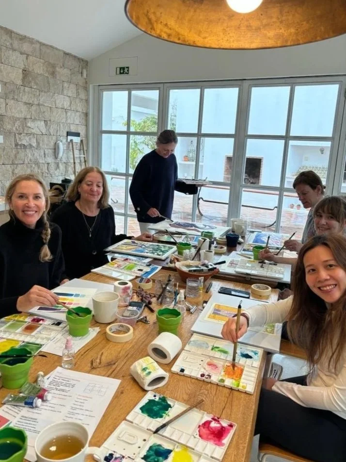 A watercolour workshop at Casa Fuzetta with founder Tara Donovan in the background — guests around the long wooden table with palettes, colour charts and paintings in progress.