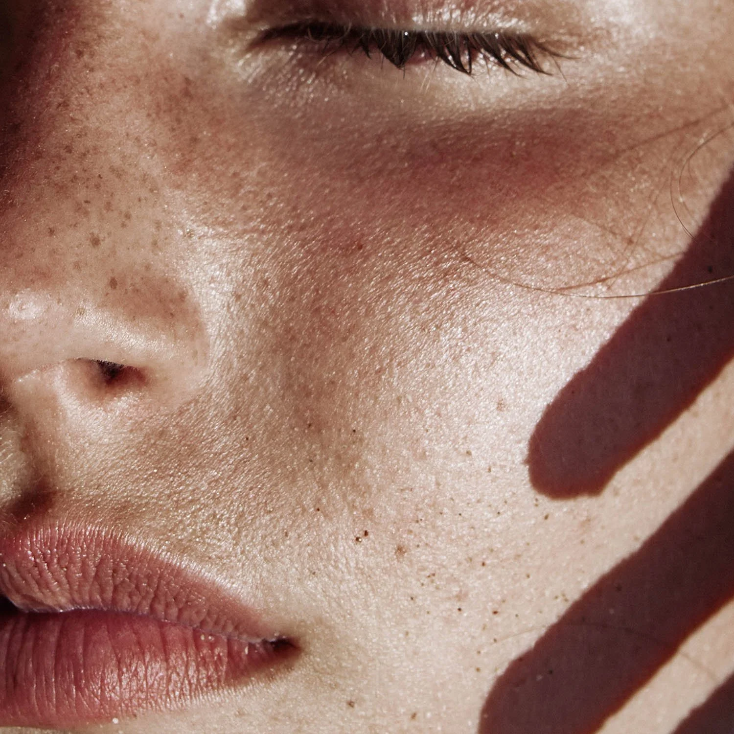 Close-up of a person's nose, lips, and part of the cheek with visible pores and skin texture, with sunlight casting a shadow on the cheek.