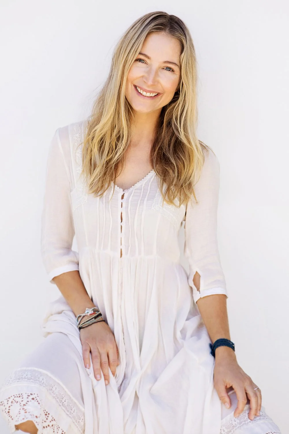 Woman with long blonde hair smiling, wearing a white dress with lace details, sitting against a plain white background.