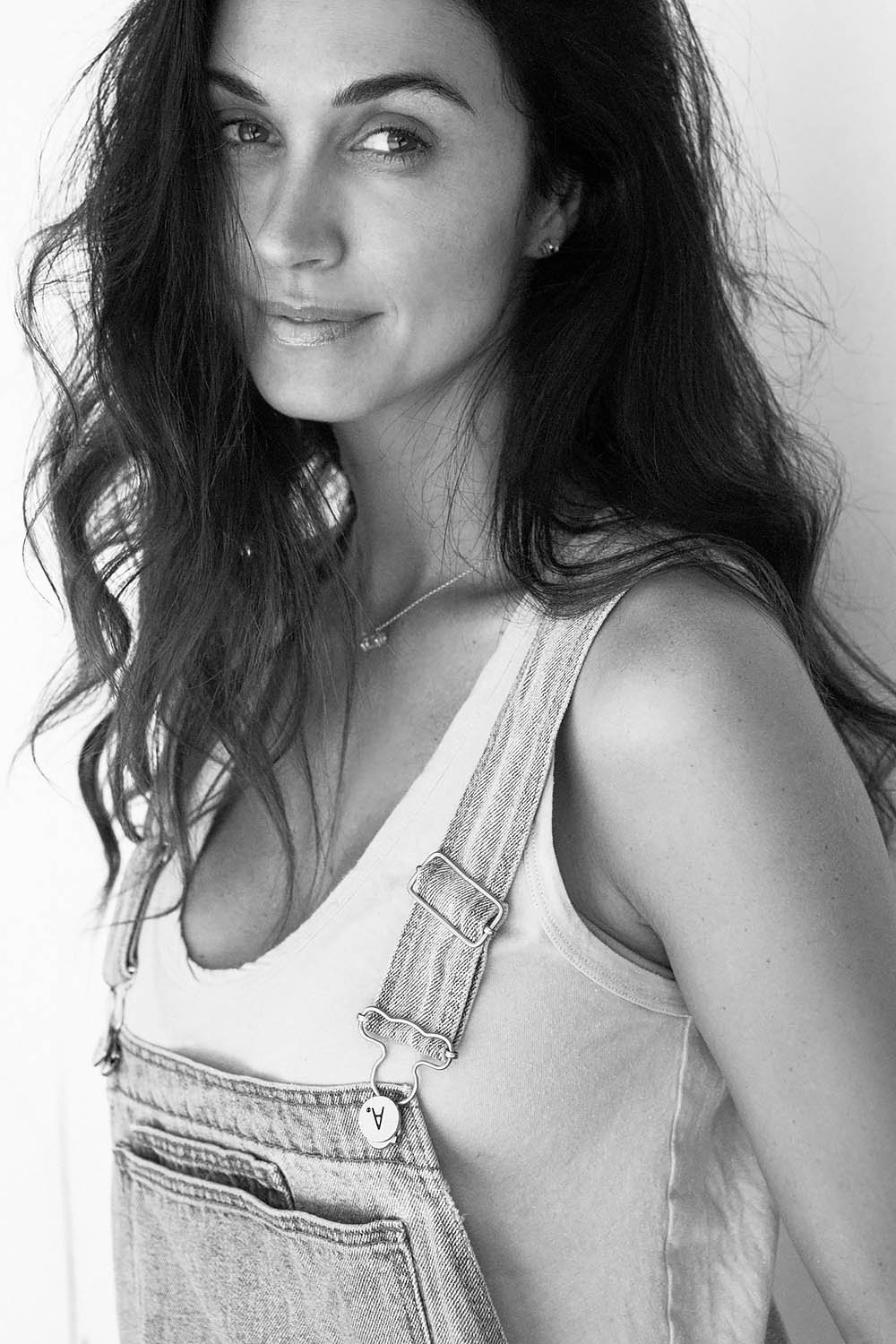 Black and white portrait of a woman with long wavy hair, wearing a light sleeveless top and denim overalls, smiling slightly and looking at the camera.