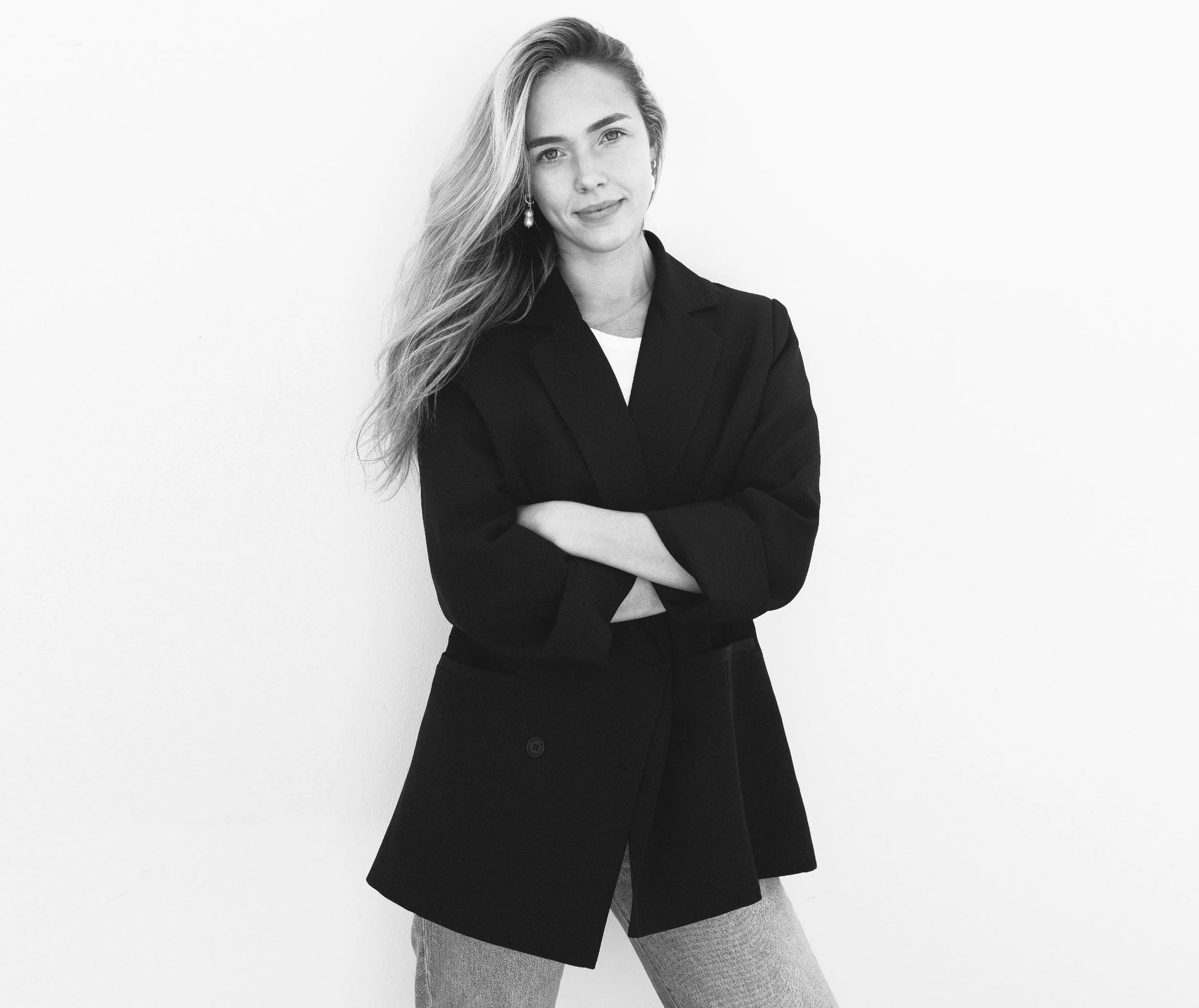 Black and white photo of a woman with long hair, crossing her arms, wearing a blazer and pants, standing against a plain background.