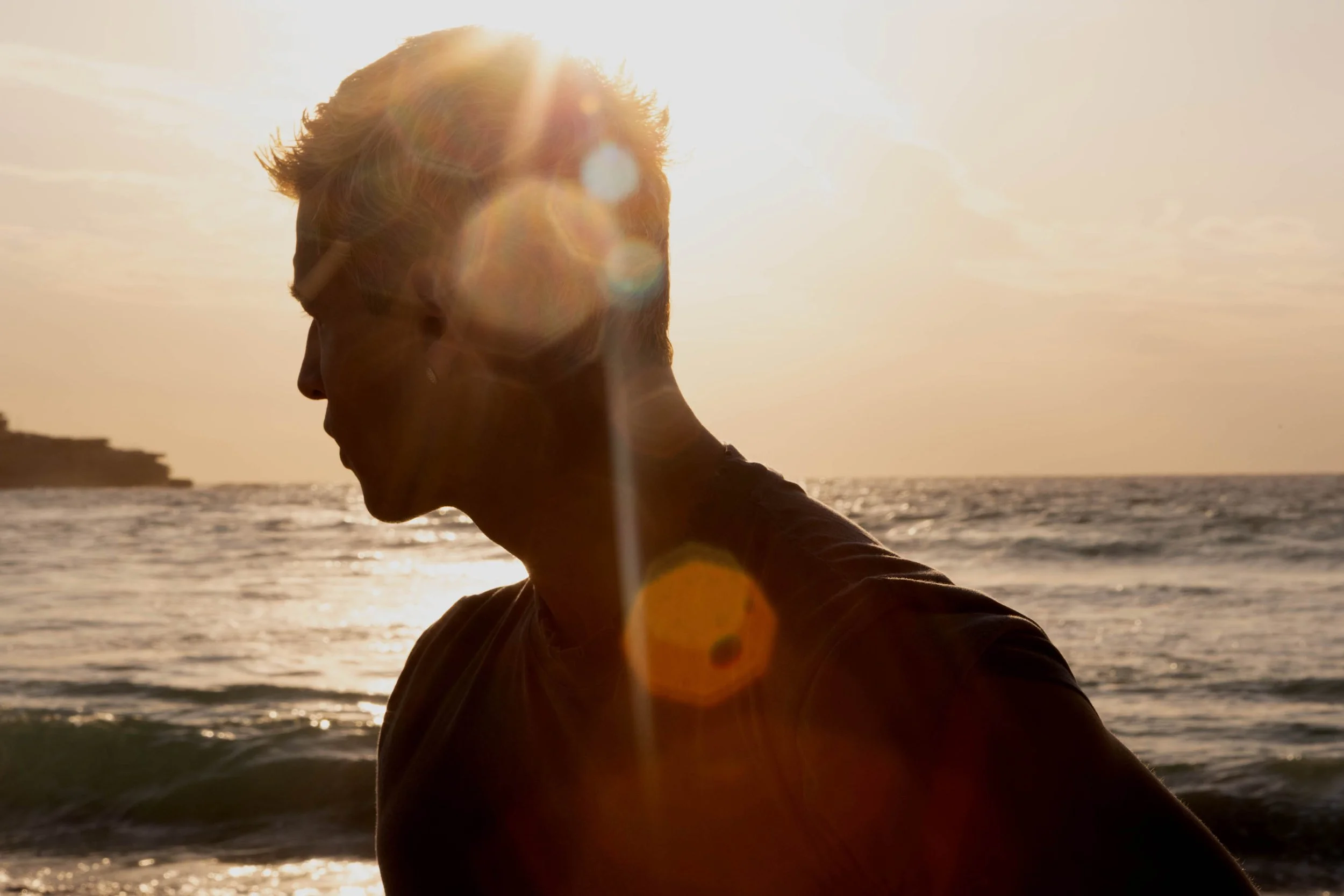 A person with short hair at the beach during sunset, with the sun shining behind their head and a lens flare effect.