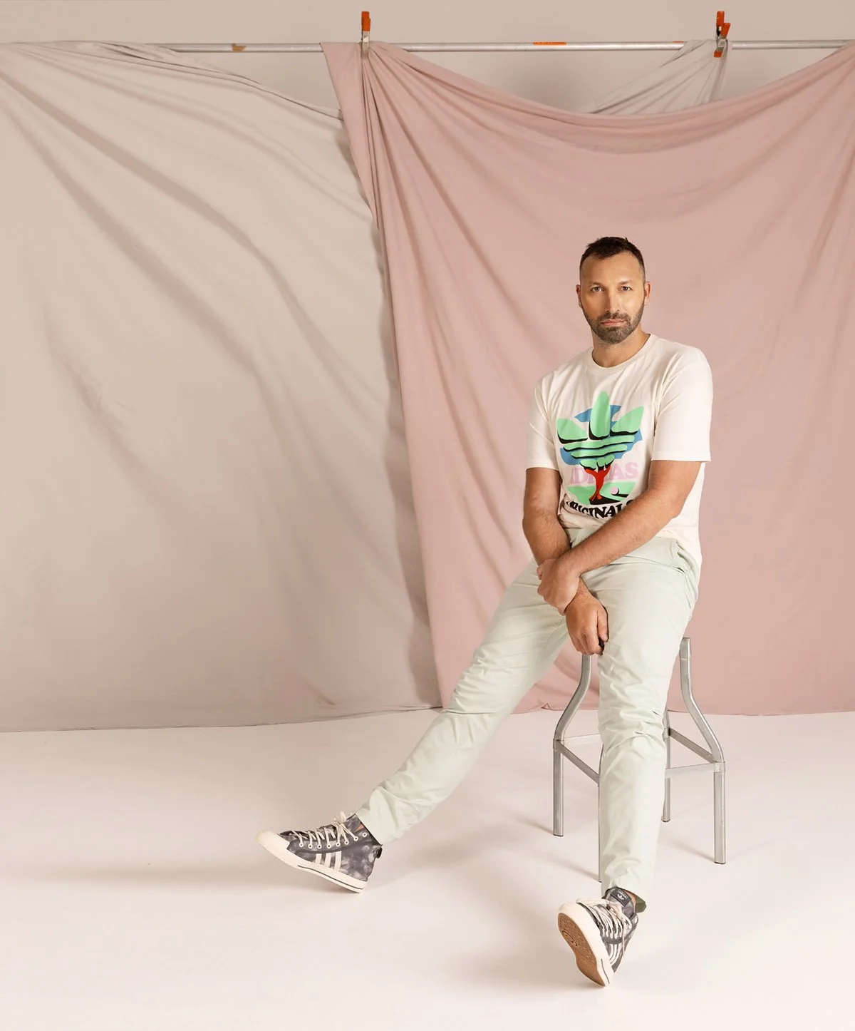 A man sitting on a stool in front of a backdrop with pink and beige fabric drapes. He is wearing a white graphic T-shirt, white pants, and patterned sneakers.