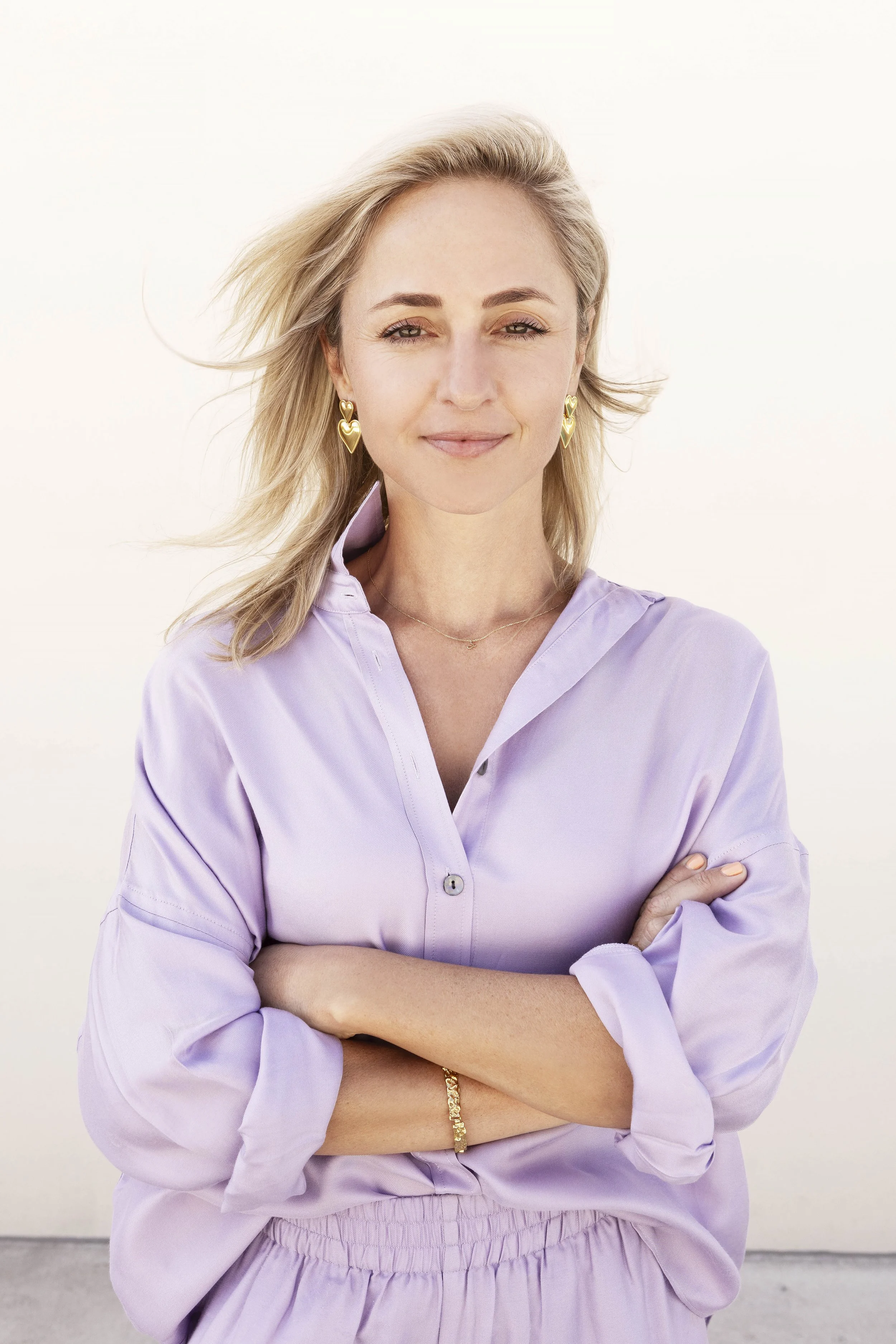 A woman with blonde hair, wearing gold heart-shaped earrings, a gold bracelet, and a light purple button-up shirt with rolled-up sleeves, standing with arms crossed against a plain light background.