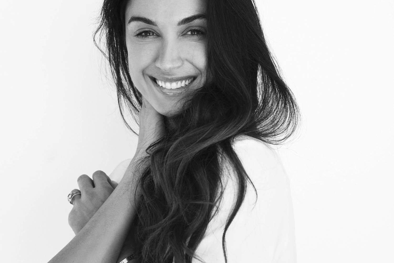 A black and white photo of a woman with long wavy hair smiling and touching her neck.