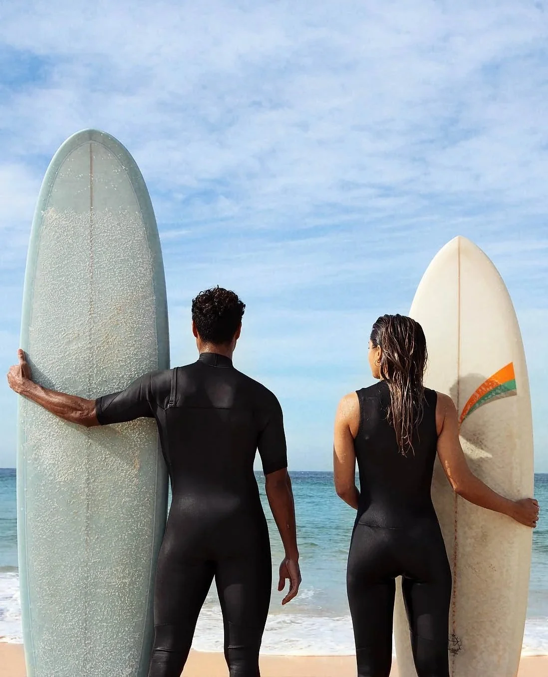 A man and a woman in black wetsuits standing on a beach, holding surfboards, facing the ocean under a partly cloudy sky.