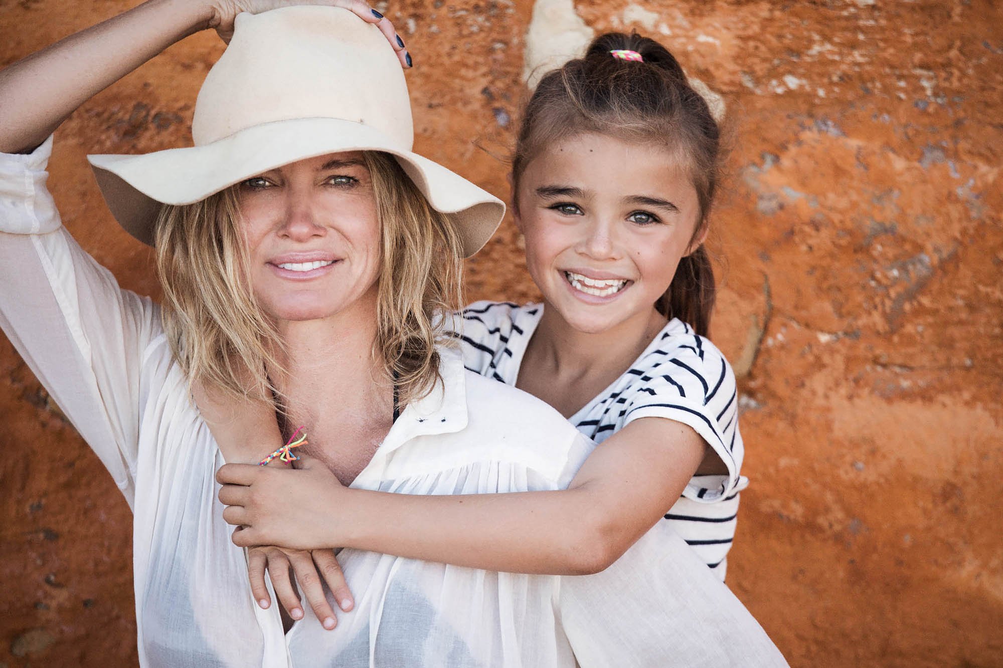 A woman with blonde hair wearing a wide-brimmed hat and a white blouse, smiling, with a young girl with brown hair in a ponytail, wearing a striped shirt, hugging her from behind against a reddish-brown rocky background.