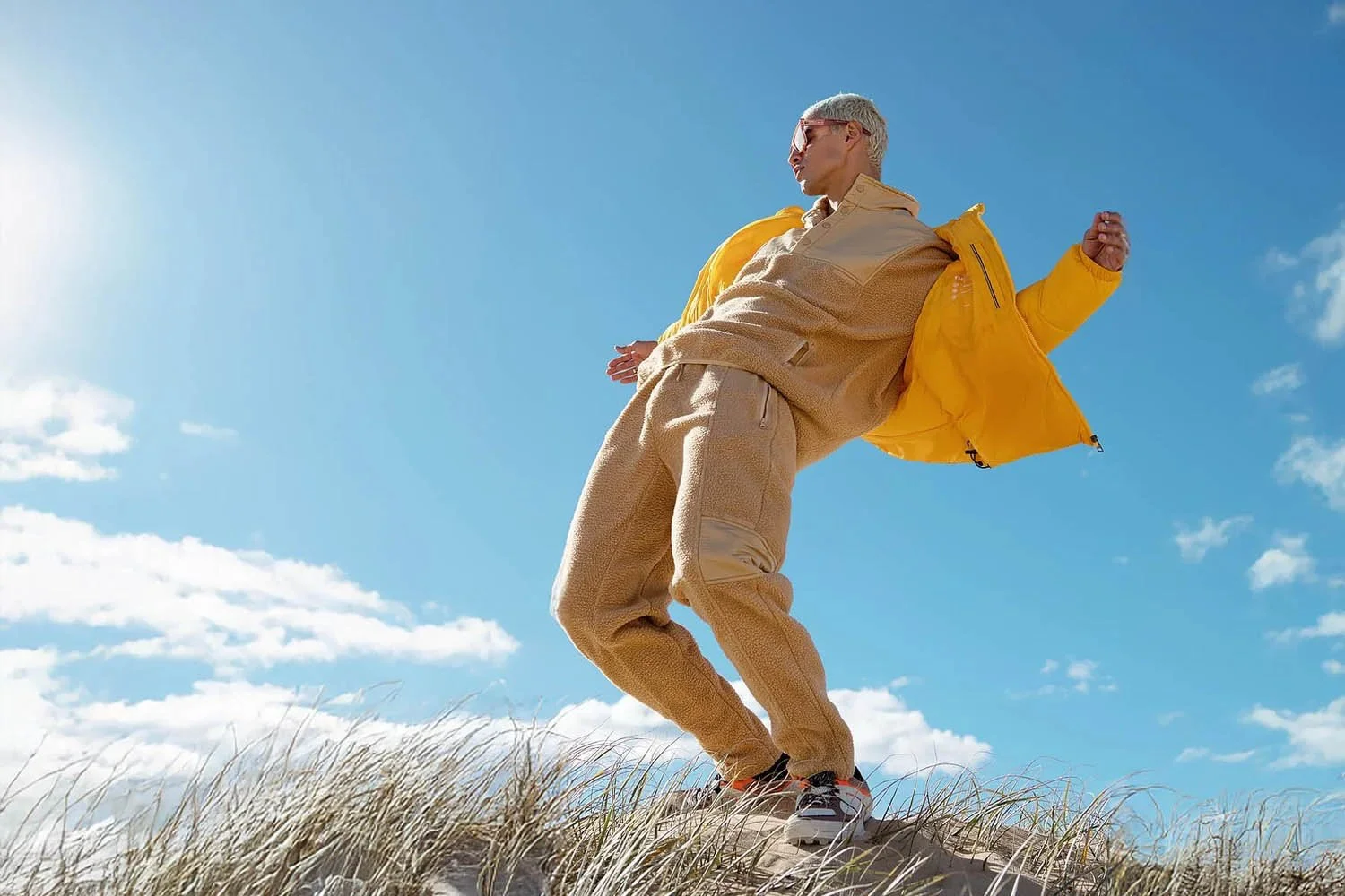 A person with blonde hair wearing sunglasses, a beige jacket, beige pants, and sneakers, standing on a sand dune against a blue sky with some clouds.