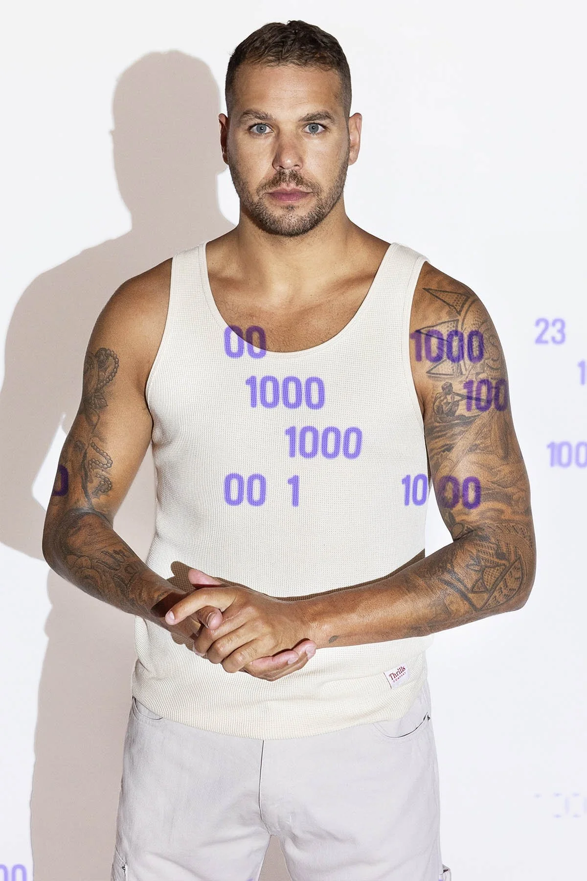 A man with tattoos wearing a white tank top and light-colored shorts, standing against a plain background with purple text and numbers overlayed, casting a shadow on the wall.