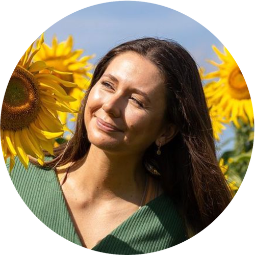 A woman with long dark hair standing in a sunflower field, smiling and looking to the side, under a blue sky with some clouds.