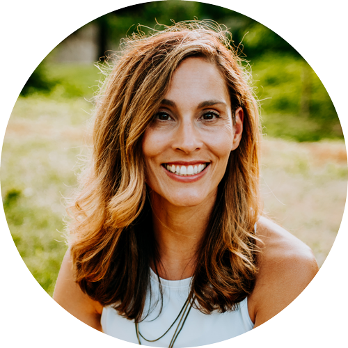 A woman with wavy, shoulder-length brown hair smiling outdoors with a blurred green background.