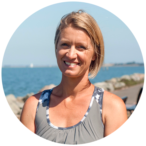 A woman with short blonde hair smiling outdoors near a body of water, wearing a sleeveless athletic top.