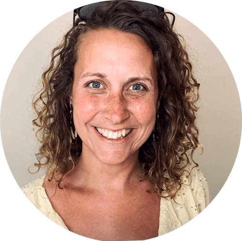 A woman with curly brown hair, fair skin, and blue eyes smiling at the camera, wearing earrings and sunglasses on her head.