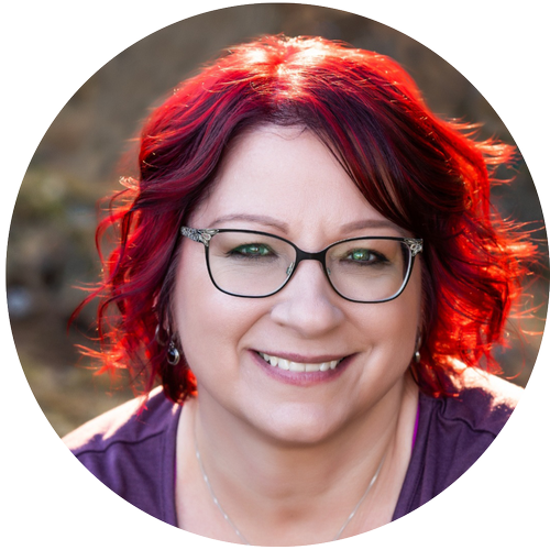 A woman with red hair, glasses, and a purple top smiling outdoors with a blurred background.