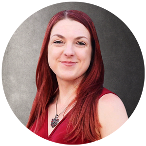 A woman with long red hair wearing a sleeveless red top and a heart-shaped necklace, smiling against a neutral background.