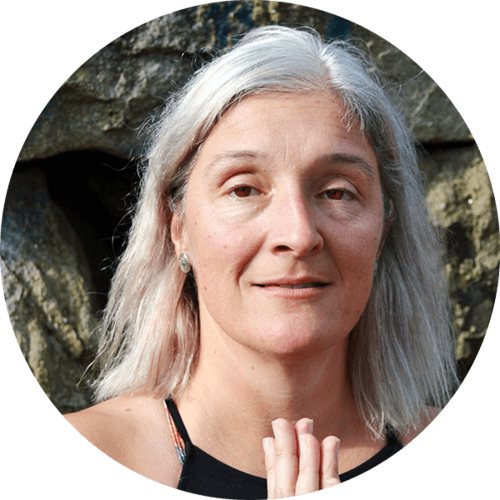 A middle-aged woman with long gray hair, holding her hands together, standing outdoors with large rocks in the background.