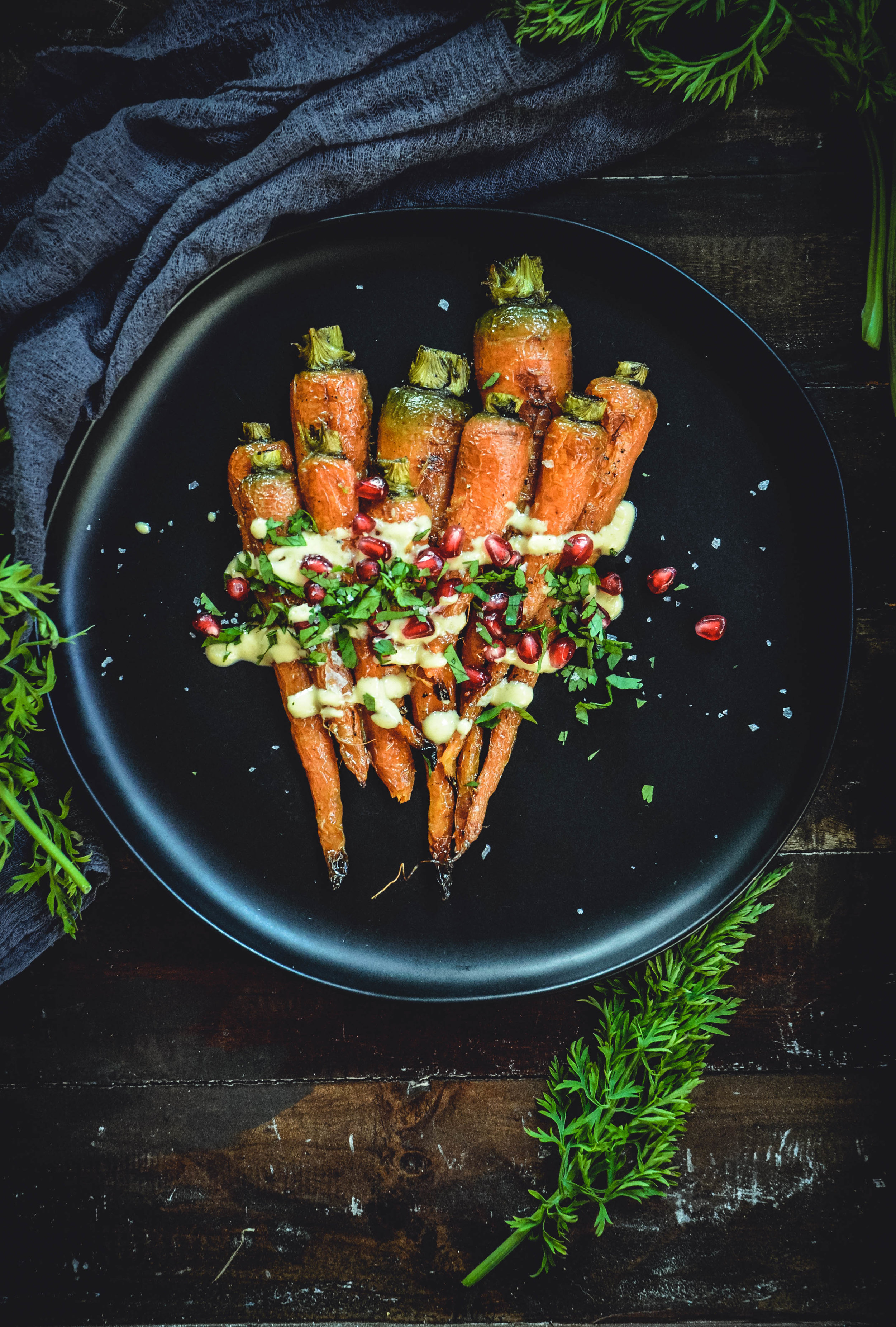 Roasted Carrots With A Simple Orange Tahini Sauce And Pomegranate on black plate with napkin and carrot greens